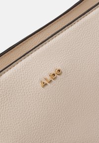 Beige texturovaná kožená taška se zlatým logem "ALDO". Má hladký vnitřní povrch a kontrastní černé obšití podél okrajů.