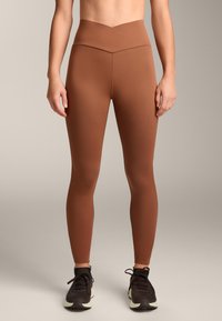 Barna, magas derekú leggings sima, rugalmas anyagból, V-alakú derékpánttal, fekete sportcipőkkel párosítva.