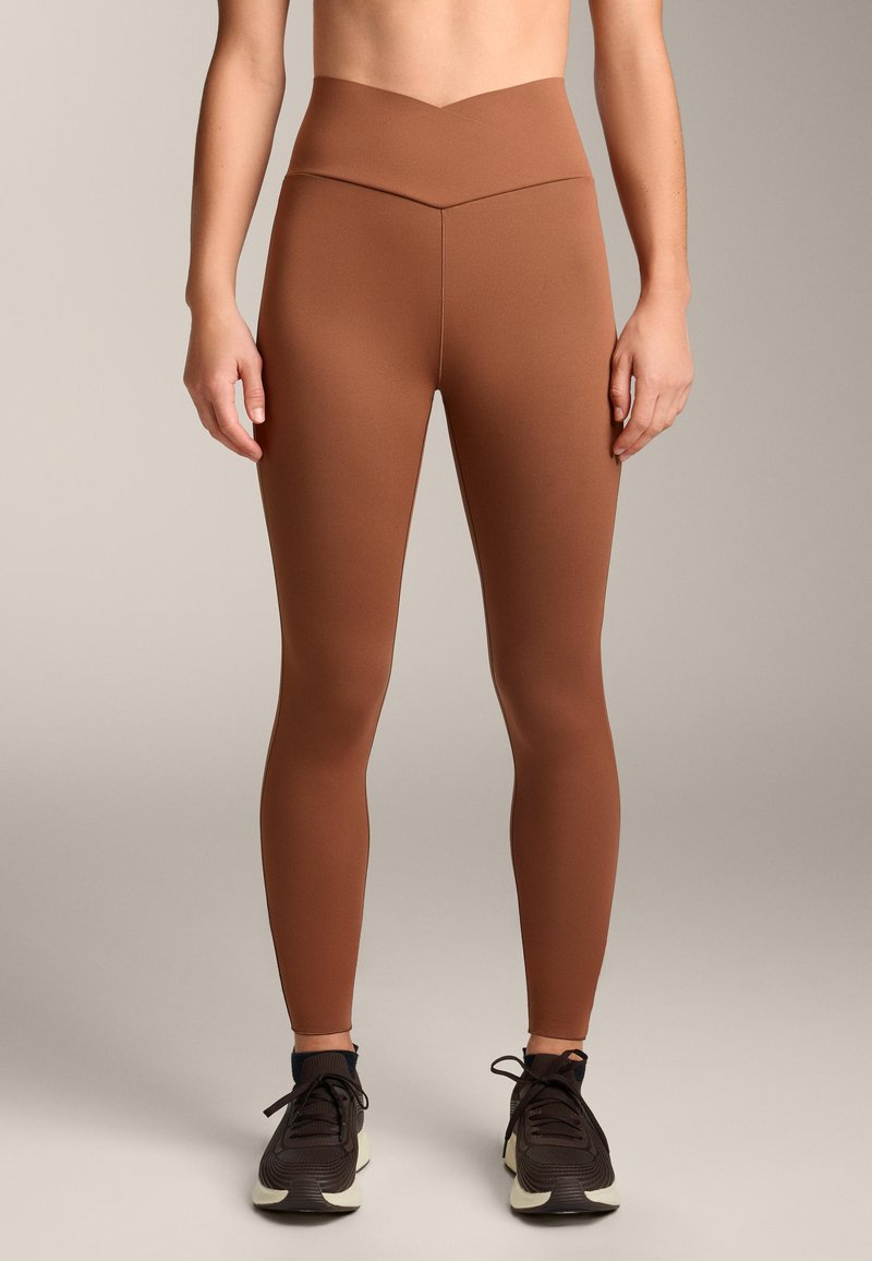Barna, magas derekú leggings sima, rugalmas anyagból, V-alakú derékpánttal, fekete sportcipőkkel párosítva.