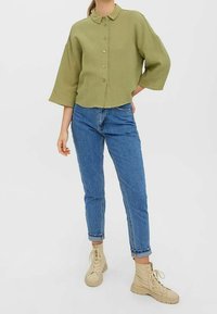 Short green button-up shirt with a textured material, wide sleeves, paired with blue jeans and beige lace-up boots.