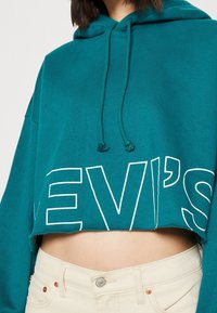 Hoodie court bleu sarcelle avec logo "LEVI’S" blanc, doté d'une capuche à cordon de serrage et d'une coupe décontractée, associé à un pantalon beige clair.