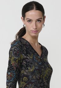Mujer con el cabello oscuro en una coleta que lleva pendientes de aro plateados y una blusa negra con un estampado floral y un cuello con cremallera.