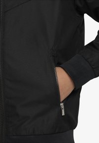 Hand insatt i sidofickan med dragkedja på svart jacka med ribbad mudd och fåll, som visar tygtextur och dragkedjedetalj.