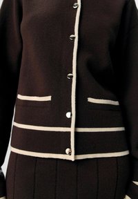 Brun stickad cardigan med vita randiga detaljer, utrustad med silverfärgade knappar, två framfickor och en texturerad tyg.