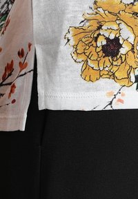 Biała koszula z żółtym kwiatowym nadrukiem po boku oraz czarnym materiałem na dole. Koszula ma krótkie rękawy i gładką fakturę.