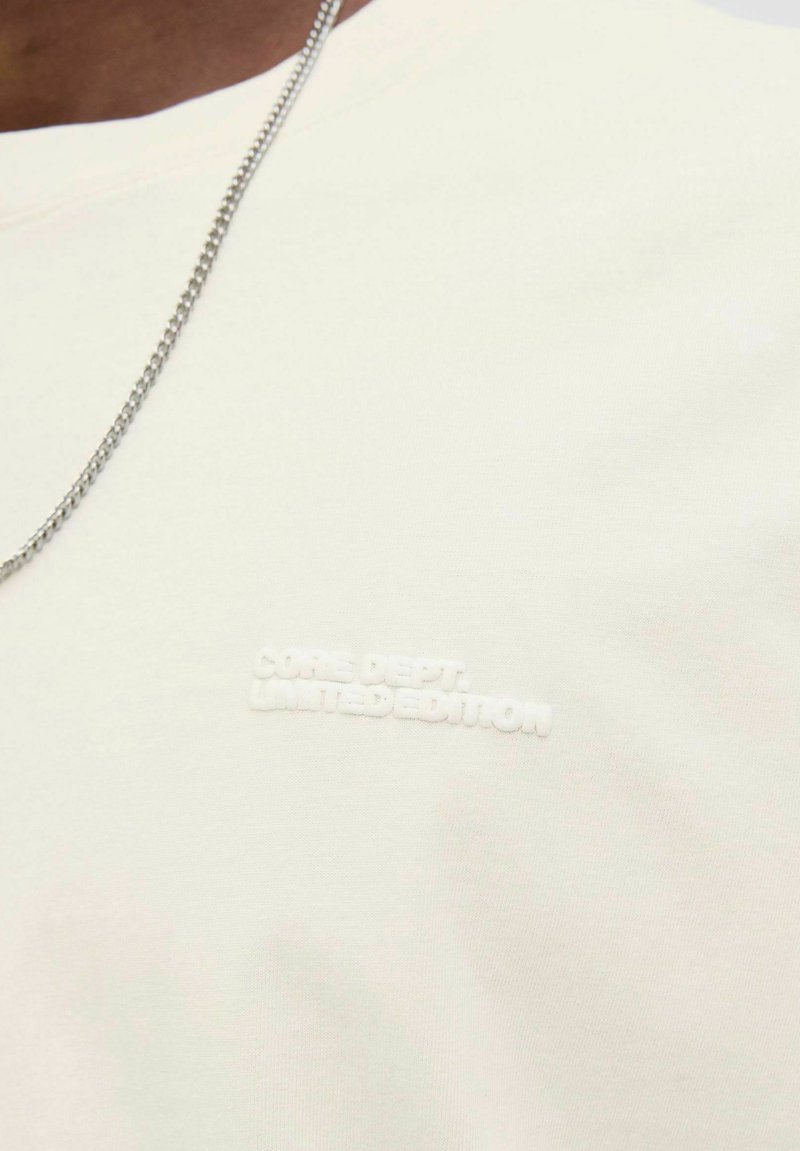 Camiseta de algodón blanca con logo texturizado "CORE DEPT. LIMITED EDITION", mangas cortas y diseño de cuello redondo. Cadena de metal visible.