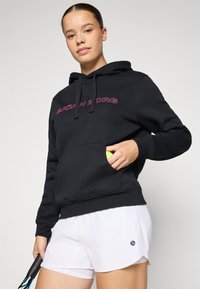 Hoodie preto feito de tecido macio, com um bolso frontal, apresentando texto de logótipo em cor rosa. Combinado com calções brancos e segurando uma raquete e uma bola de ténis.