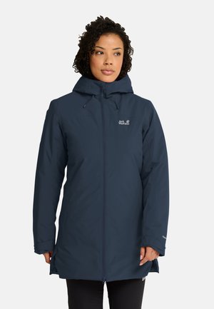 Vrouw draagt een lange donkerblauwe winterjas met capuchon, helemaal dichtgeritst, staand met ontspannen handen langs het lichaam tegen een effen achtergrond.