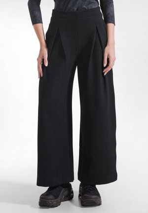 Personne portant un pantalon noir taille haute à jambes larges avec des plis, associé à des baskets noires épaisses, debout devant un fond blanc uni.