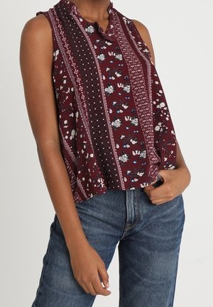 Mouwloze wijnrode blouse met verticale bloem- en geometrische patronen, gecombineerd met een blauwe spijkerbroek, gedragen door een staand persoon met één hand in de zak.