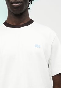 Gros plan d'une personne portant un t-shirt blanc avec un col noir et un petit logo de crocodile bleu clair sur la poitrine.