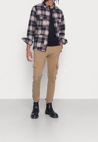 Černobílá kostkovaná košile jako bunda se dvěma předními kapsami, kombinovaná s béžovými cargo kalhotami se dvěma bočními kapsami a černými kotníkovými botami.