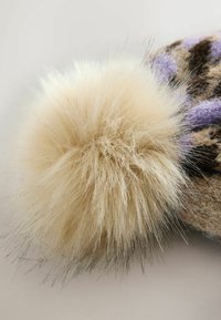 Faux-Fur-Pompon in cremigem Beige, flauschiger Textur, mit feinen dunkleren Strähnen, befestigt an einer gemusterten Wollbasis mit braunen und lila Farbtönen.
