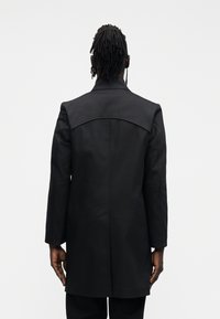 Czarny płaszcz z wysokim kołnierzem i usztywnionymi ramionami. Gładka tkanina o prostym kroju, sięgająca do połowy uda, z długimi rękawami.