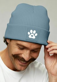 Smiling man adjusting light blue knit beanie with white paw print and heart emblem, wearing white shirt, neutral background.