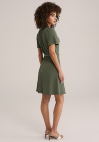 Kortärmad grön klänning med texturerad tyg, figursydd vid midjan och med en utsvängd kjol. Vita sandaler kompletterar outfiten.