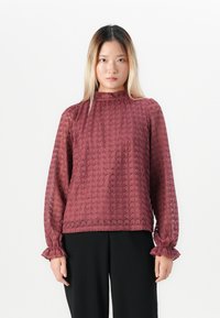 Vrouw met steil haar in een lange mouw rozenkant blouse met ruffles aan de manchetten en zwarte broek, staand tegen een witte achtergrond.