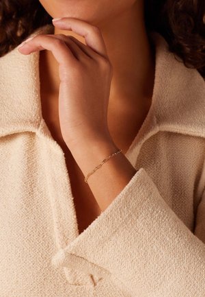 Bracelet en chaîne dorée sur un poignet, arborant des formes ornées entrelacées. La main est proche d'un tissu texturé beige clair avec de grands revers.