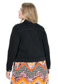 Svart denimjacka med silverfärgade nitar vid axlarna och ärmarna, bärs över en mönstrad, flerfärgad klänning.