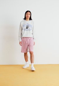 Gray sweatshirt with blue text and graphic, paired with pink shorts. Model wearing white sneakers and ankle socks, standing on yellow floor.