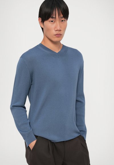Μπλε ριγέ μακρυμάνικο πουλόβερ με σχέδιο V-λαιμού, υφή υφάσματος και εφαρμοστή σιλουέτα, σε συνδυασμό με σκούρες παντελόνες.