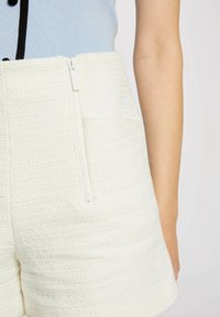 I pantaloni corti bianchi testurizzati presentano una chiusura con zip frontale e una tasca laterale. Il tessuto ha un aspetto morbido e intrecciato, accentuando un design strutturato.