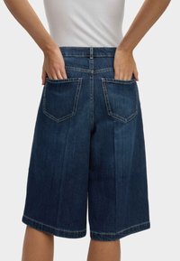 Dunkelblaue Denim-Culottes mit einem entspannten Schnitt, zwei Gesäßtaschen, sichtbaren Nähten und einem geraden Saum, der unterhalb des Knies endet.