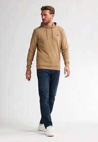 Felpa beige con cappuccio e logo, abbinata a jeans blu scuro e sneaker bianche. Il tessuto sembra liscio con una vestibilità comoda.