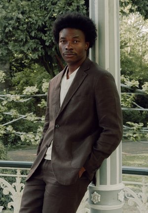 Mann mit Afro-Frisur, der einen braunen Anzug und ein weißes Hemd trägt und sich an eine Säule in einem blattreichen Außenbereich lehnt.