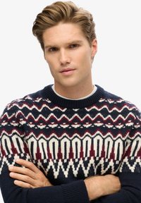 Marineblauer Wollpullover mit geometrischem Muster in Creme und Weinrot. Rundhalsausschnitt, strukturstrick, mit langen Ärmeln und gerippten Bündchen.