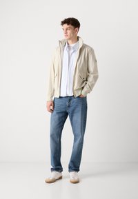 Lys beige bomberjakke med ståkrage, glidelås og sidelommer, kombinert med en blå skjorte med knapper og blå jeans. Hvite sneakers.