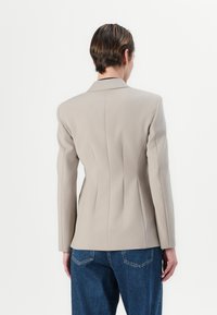Beige vypasovaný blazer se strukturovanými rameny, vertikálními švy a jedním zadním rozparkem, spárovaný s volnými modrými džínami.