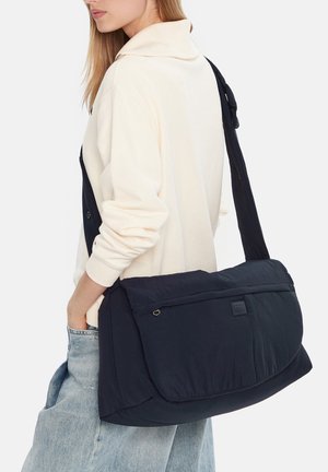 Žena v světle béžovém svetru a světle modrých džínách nese velkou černou polstrovanou tašku přes rameno na zádech.