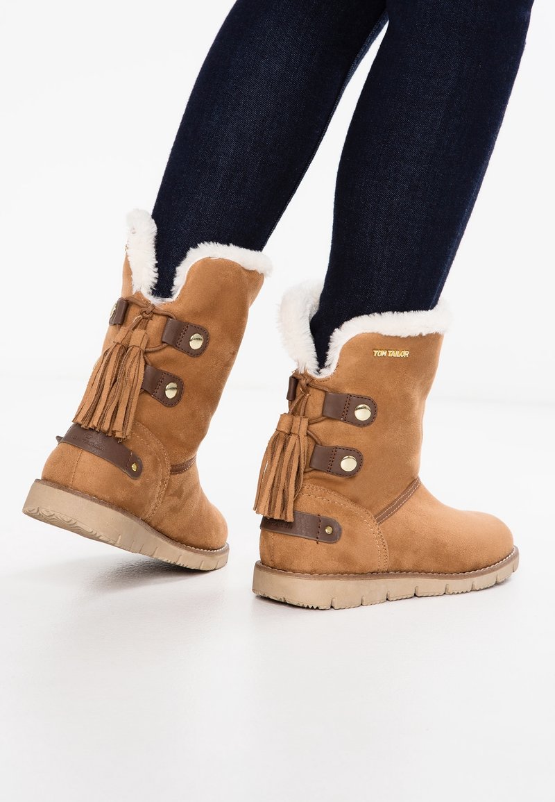 Bottines en suède marron avec bordure en fourrure blanche, pampilles décoratives et accents en cuir marron. Semelles plates avec une texture antidérapante.