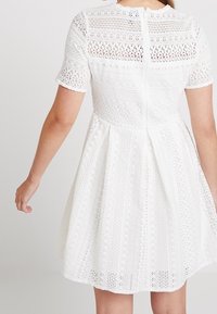 Biała sukienka z koronką, z krótkimi rękawami, z misternymi wzorami, obcisłym gorsetem i rozkloszowanym dołem. Widoczny zamek błyskawiczny z tyłu.