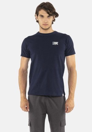 Jeune homme debout portant un t-shirt bleu marine Leone 1947 et un pantalon cargo gris foncé sur un fond clair uni.