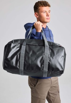 Joven con sudadera azul con capucha que lleva una bolsa de deporte negra grande sobre el hombro frente a un fondo gris liso.