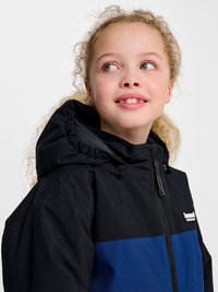 Jong meisje met krullend blond haar dat een zwart-blauwe capuchonjas draagt, kijkt naar rechts met een lichte glimlach tegen een witte achtergrond.