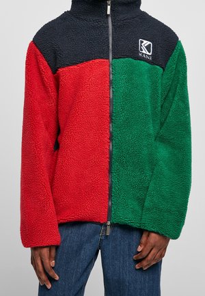 Homme portant une veste polaire à fermeture éclair complète avec une poitrine noire, côté gauche rouge, côté droit vert, et logo Kani sur la poitrine, associée à un jean bleu.