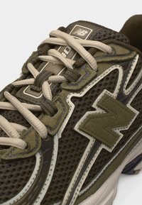 Κοντινό πλάνο σε αθλητικό παπούτσι New Balance σε ελιά πράσινο χρώμα, με δίχτυ από ύφασμα, μπεζ κορδόνια και ανασηκωμένο λογότυπο "N" στο πλάι.