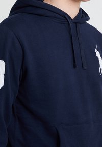 Muž oblečený v tmavě modré mikině s kapucí, na hrudi s bílým logem polo hráče a s bílým číslem na rukávu.