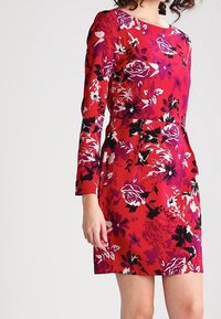 Nainen, jolla on päällään punainen kukallinen pitkähiha-l dress, jossa on purppuran, mustan ja valkoisen kukat, seisoo yksinkertaista vaaleaa taustaa vasten.