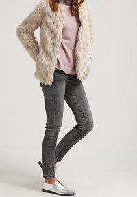 Beige fuskpälsjacka över en rosa tröja, ihop med grå smala jeans och metalliska silverskor. Texturerade material, avslappnad design.