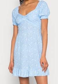 Ljusblå klänning med puffärmar och V-ringning. Har en samlad midja, blommönster och volangkant. Tillverkad av lättviktig tyg.