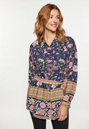 Frau lächelt, trägt eine marineblaue Bluse mit rosa und grünen Blumen und dekorativen goldenen Musterrändern, steht vor einem hellen Hintergrund.