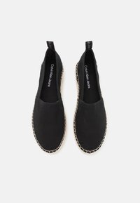 Černé plátěné nazouvací boty s texturovanou podešví, béžovým prošíváním kolem okraje a značkou na vnitřním podšívení. Bez viditelných tkaniček.