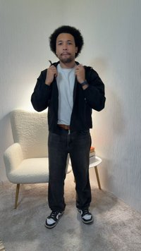 Bărbat cu păr creț, purtând o geacă neagră, cămașă albă, blugi negri și adidași, stând într-o cameră cu mochetă, lângă un scaun alb și o masă mică.