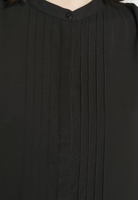 Blouse noire avec des plis verticaux, une fermeture par boutons au niveau du cou, et un tissu lisse et léger. Design simple et épuré.