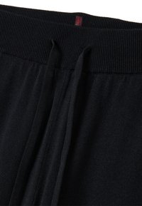 Gros plan de la ceinture de pantalon en tricot noir avec cordons de serrage et une petite étiquette rectangulaire rouge centrée sur le bandeau élastique côtelé.