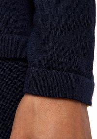 Rękaw ciemnoniebieskiego swetra z dzianiny podwinięty, odsłaniający nadgarstek i część dłoni na jasnym tle.